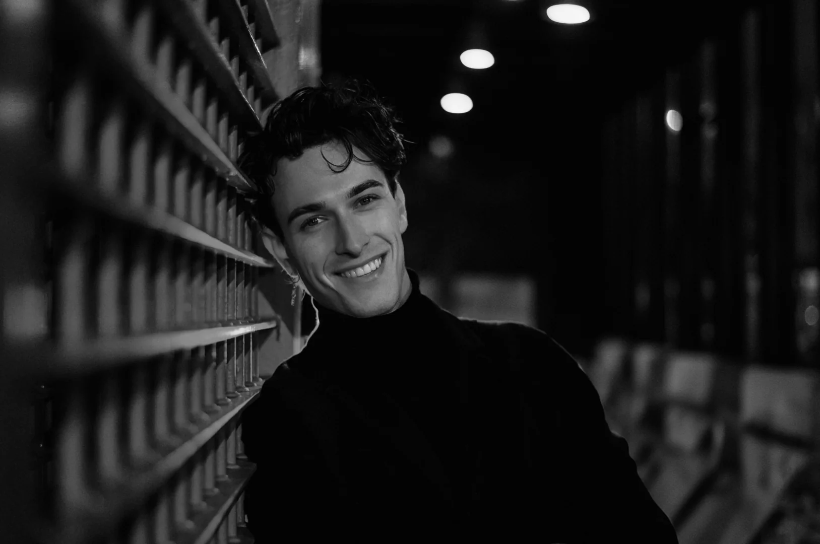Jules Sauvage, escort gay Paris — sourire noir et blanc dans une ruelle, col roulé sous guirlandes lumineuses