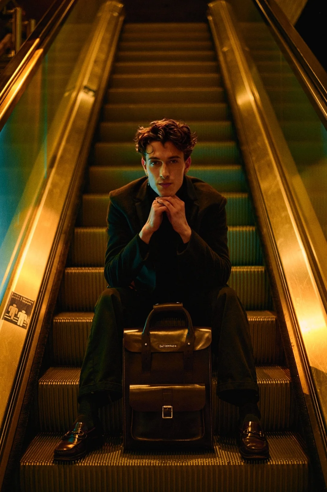Jules Sauvage, escort gay haut de gamme Paris — portrait éditorial assis sur un escalator parisien, manteau sombre, mocassins et sacoche en cuir, lumières dorées et turquoises