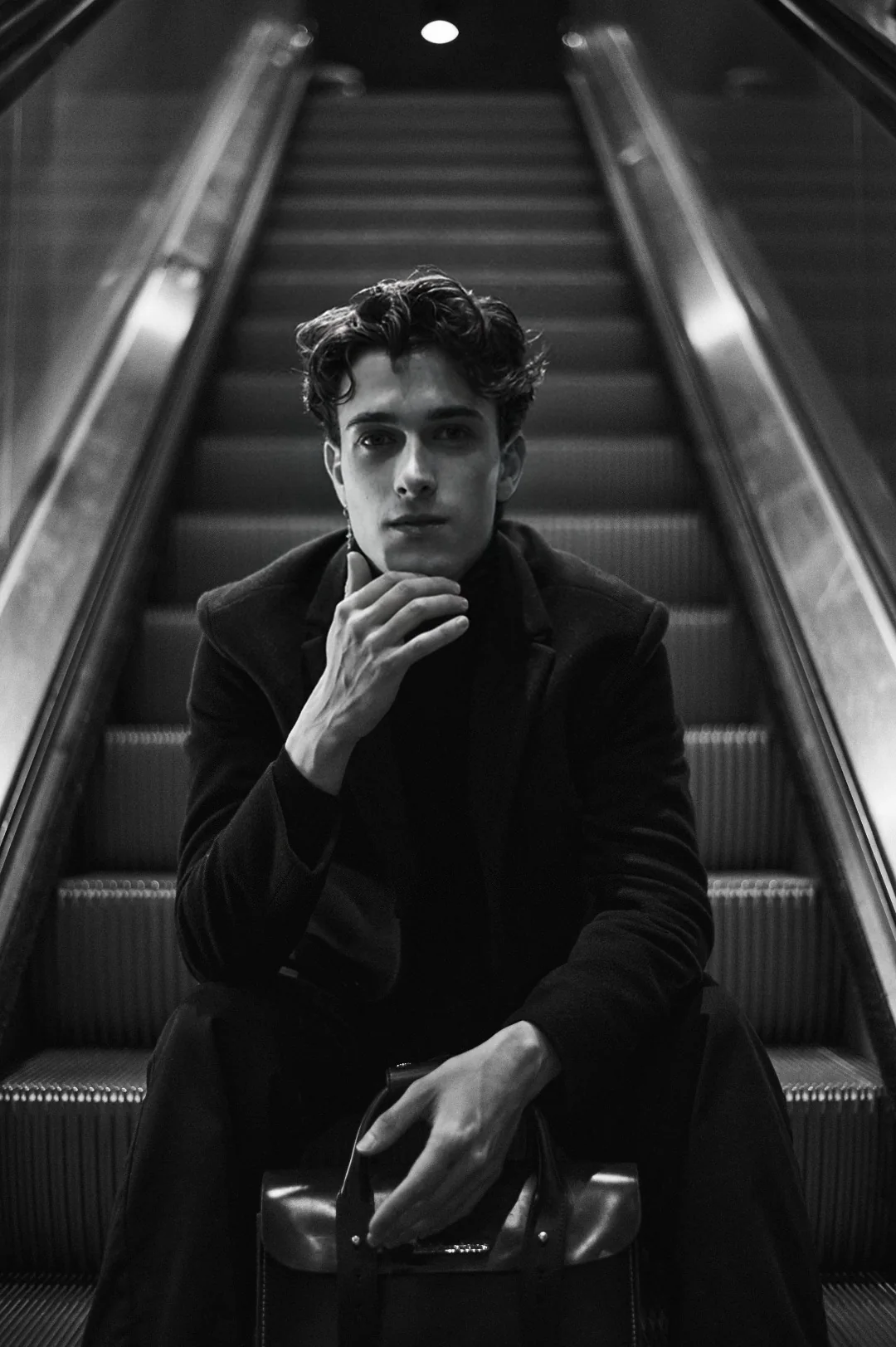 Jules Sauvage, high class gay male escort Paris — editorial black and white portrait seated on an escalator, dark coat, hands crossed on a leather satchel