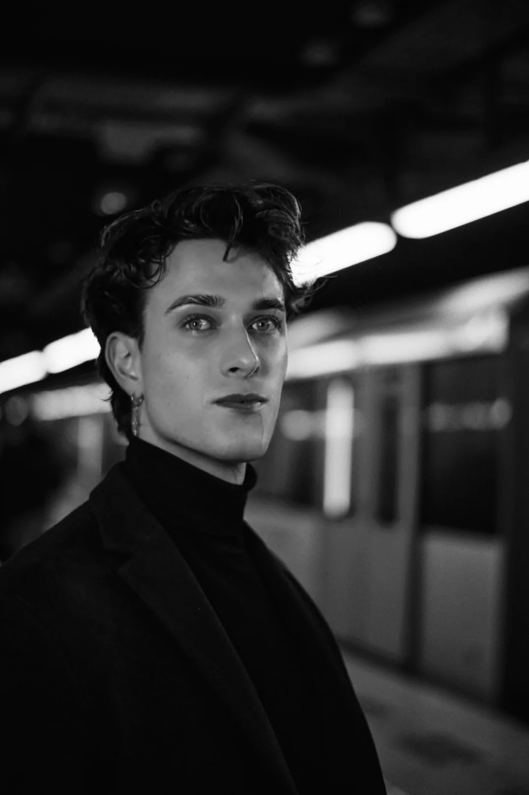 Jules Sauvage, gay male escort Paris — editorial black and white portrait in a metro station at night, black turtleneck and dark jacket